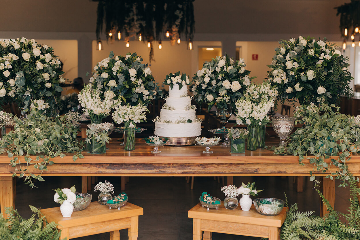 Mesa de madeira com bolo de casamento com quatro andares ao lado de arranjos com flores brancas