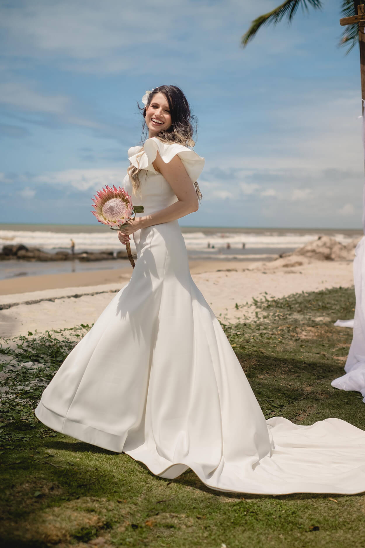 Mulher com vestido de noiva com cauda e segurando protea na praia