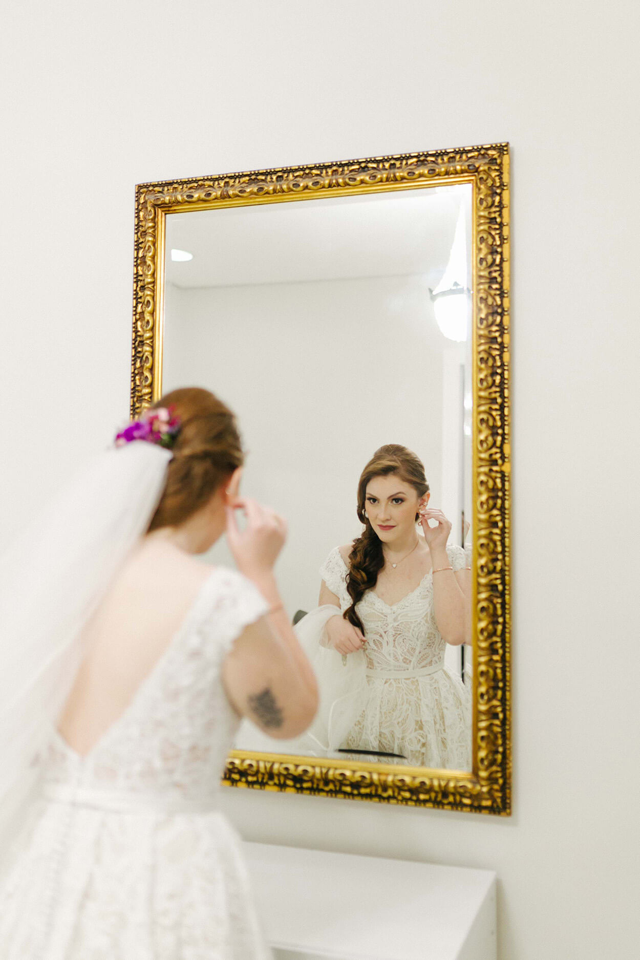 Mulher ruiva com vestido de noiva e véu se olhando no espelho com moldura clássica dourada