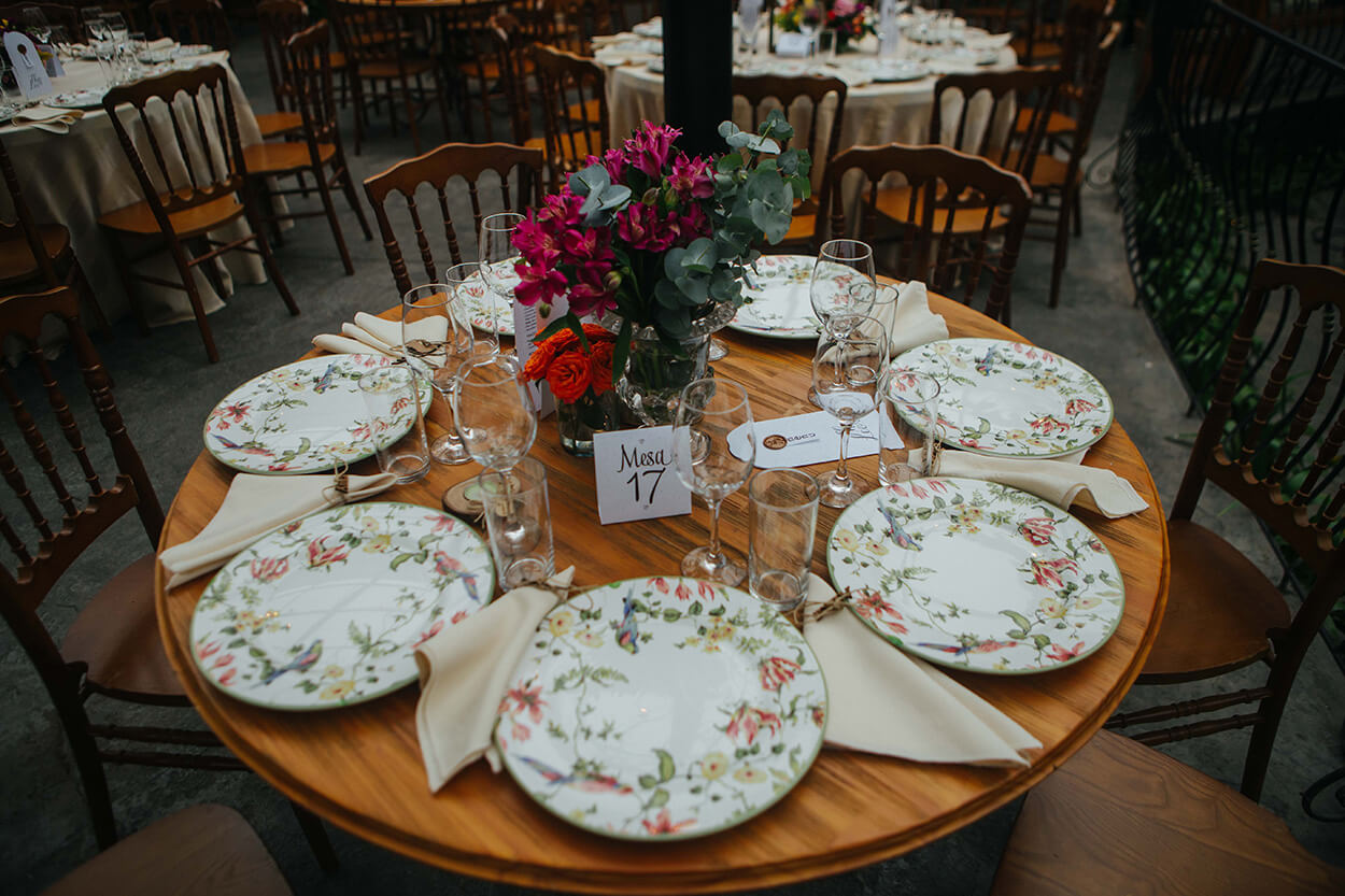 Mesa de madeira redonda posta com pratos com estampas de flores e passarinhos