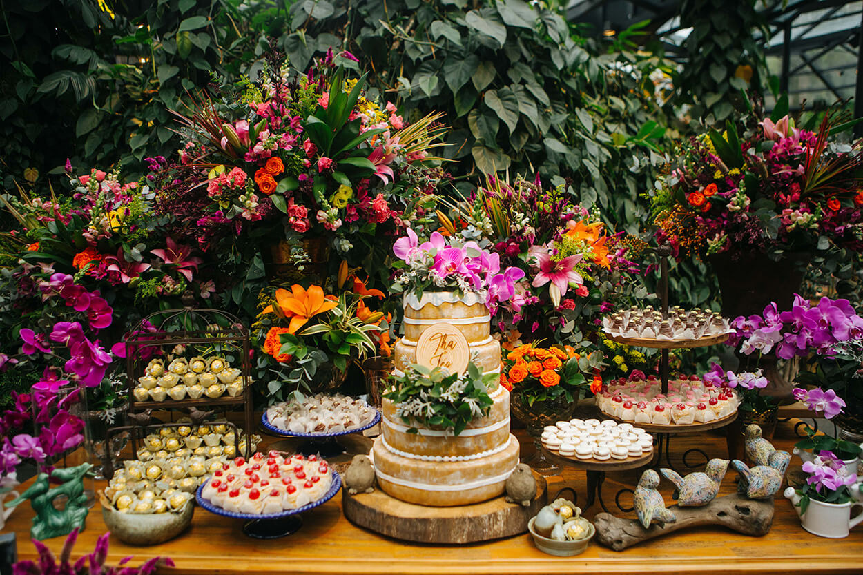 MEsa de madeira com bolo de casamento de tres andares com orqideas no topo e bandejas com doces de casamento