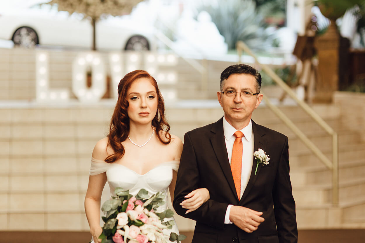 Entrada da noiva com buquê com flores rosas e brancas ao lado do pai com tenro preto e gravata laranja