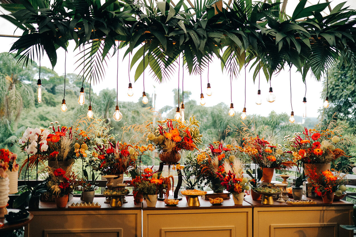 Mesa com arranjos com flores vermelhas e laranjas e arranjo pendente com folhagens e lampadas