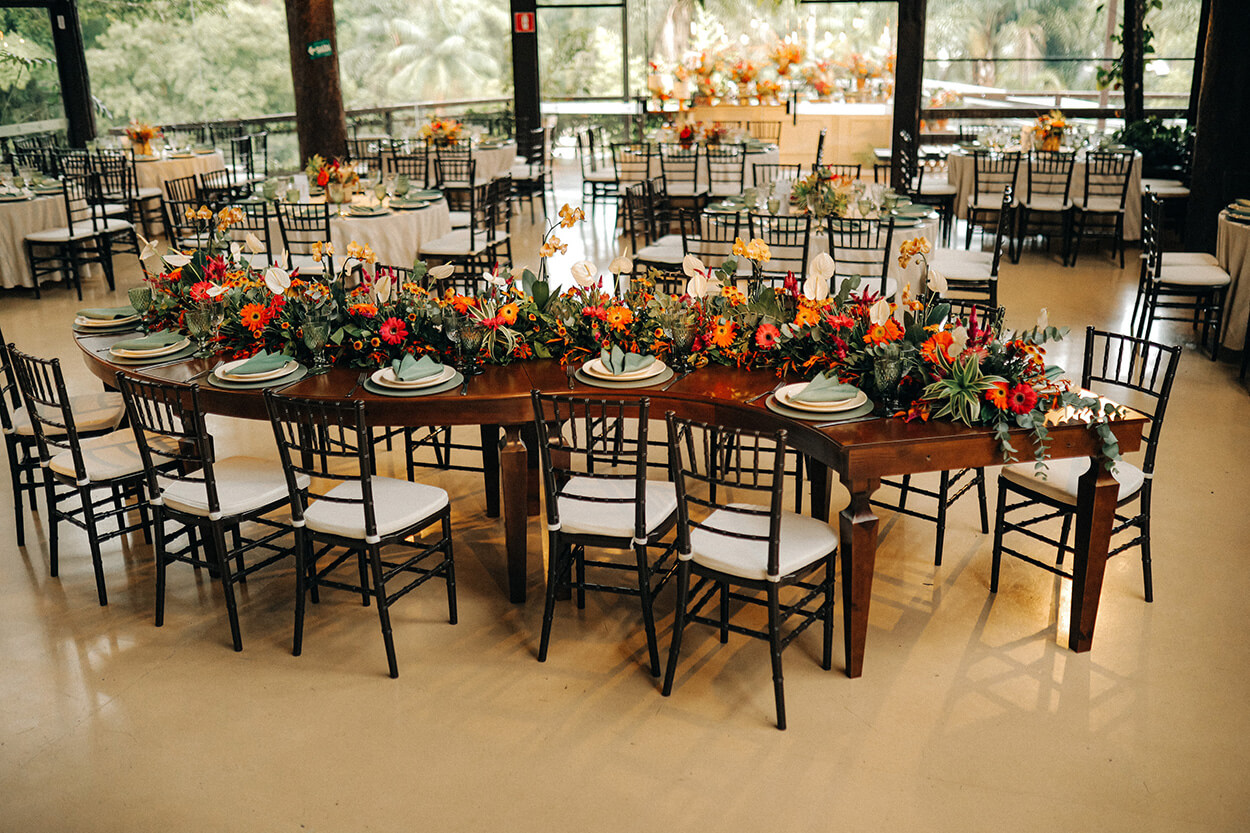 Salão com mesas postas com flores amarelas laranjas e vermelhas