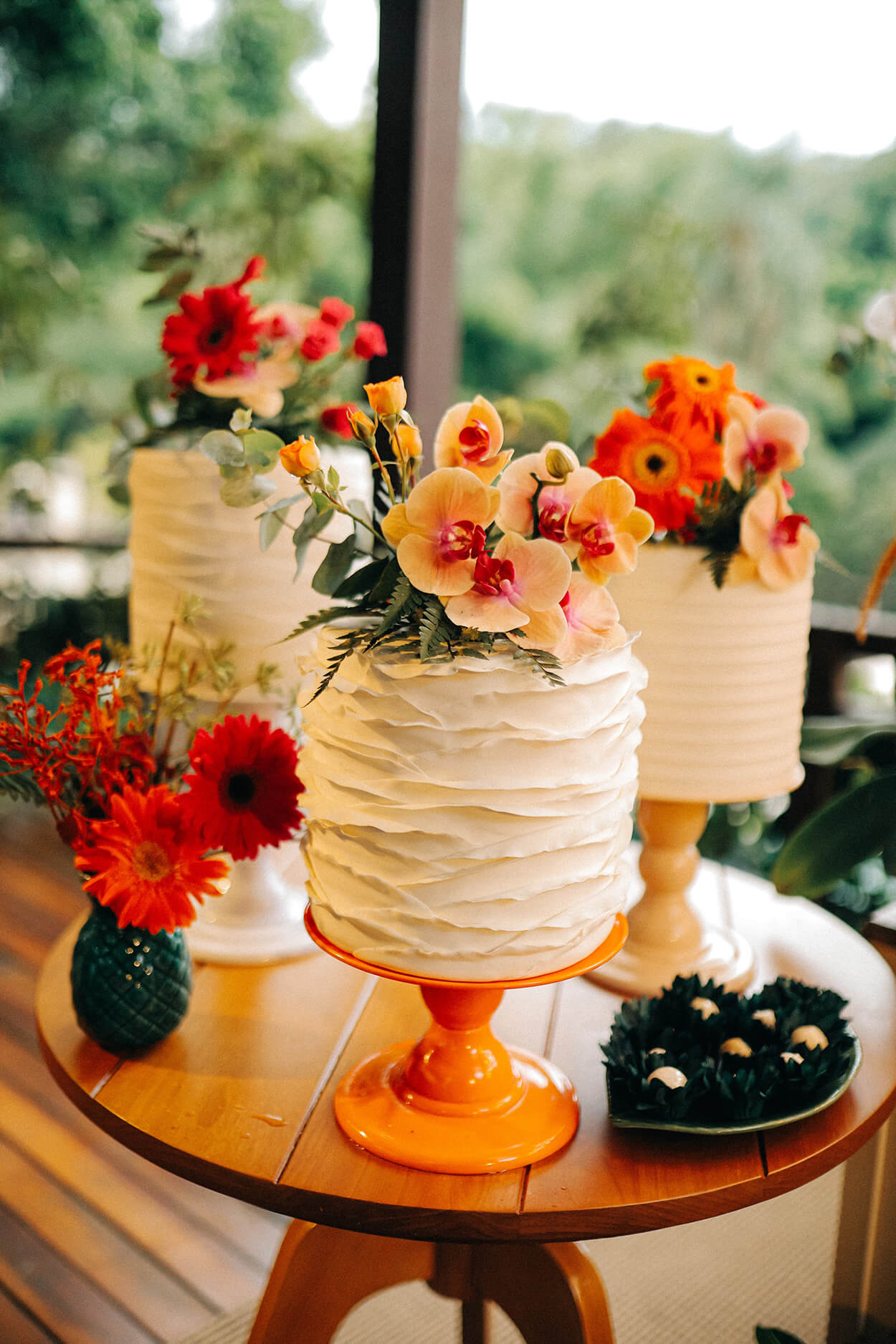Mesa de madeira redonda com três bolos de casamento com flores amarelas vermelhas e laranjas no topo