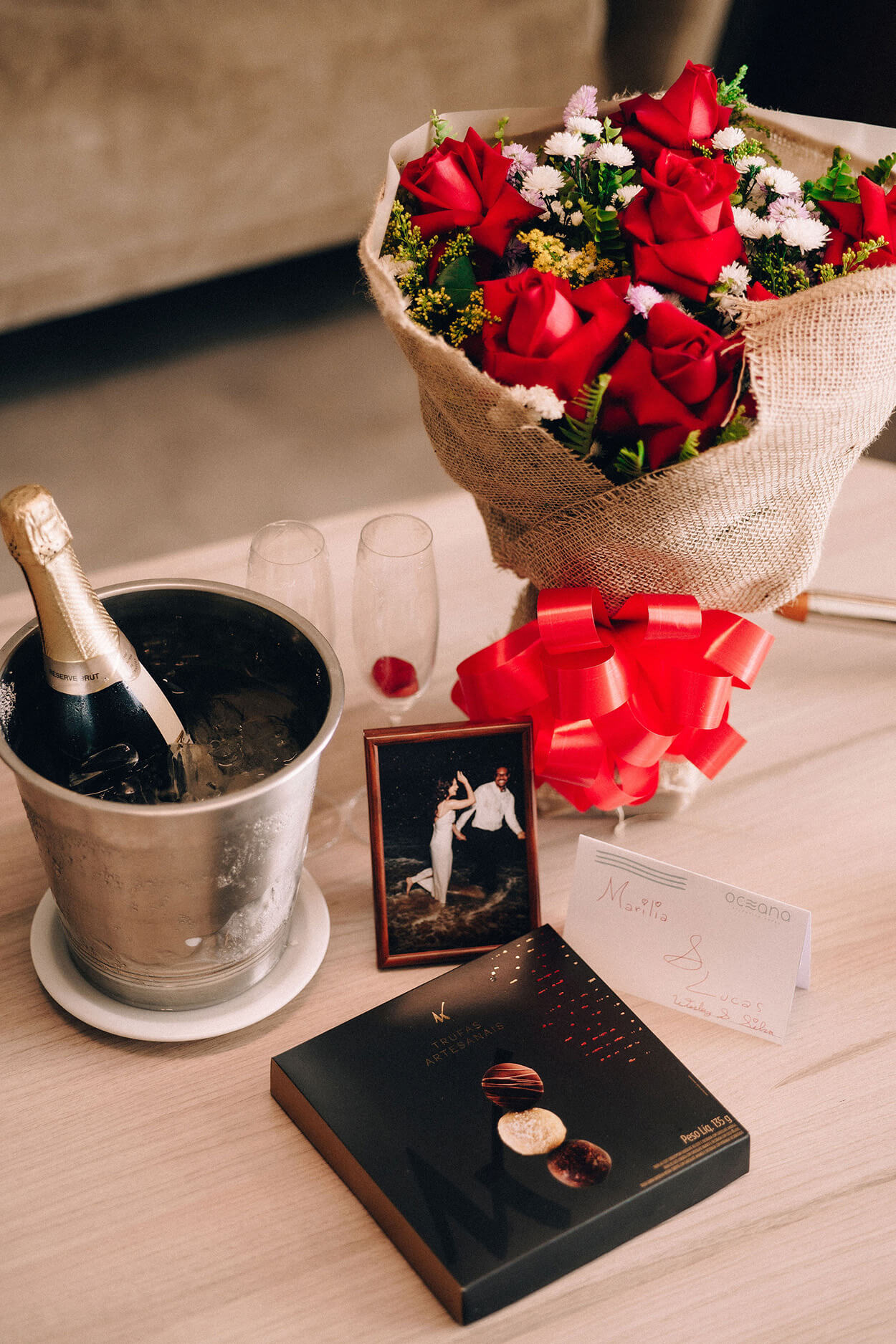 Balde com bebida no gelo porta retrao com foto do casal caixa de chocolate e buque com rosas vermelhas em cima da mesa