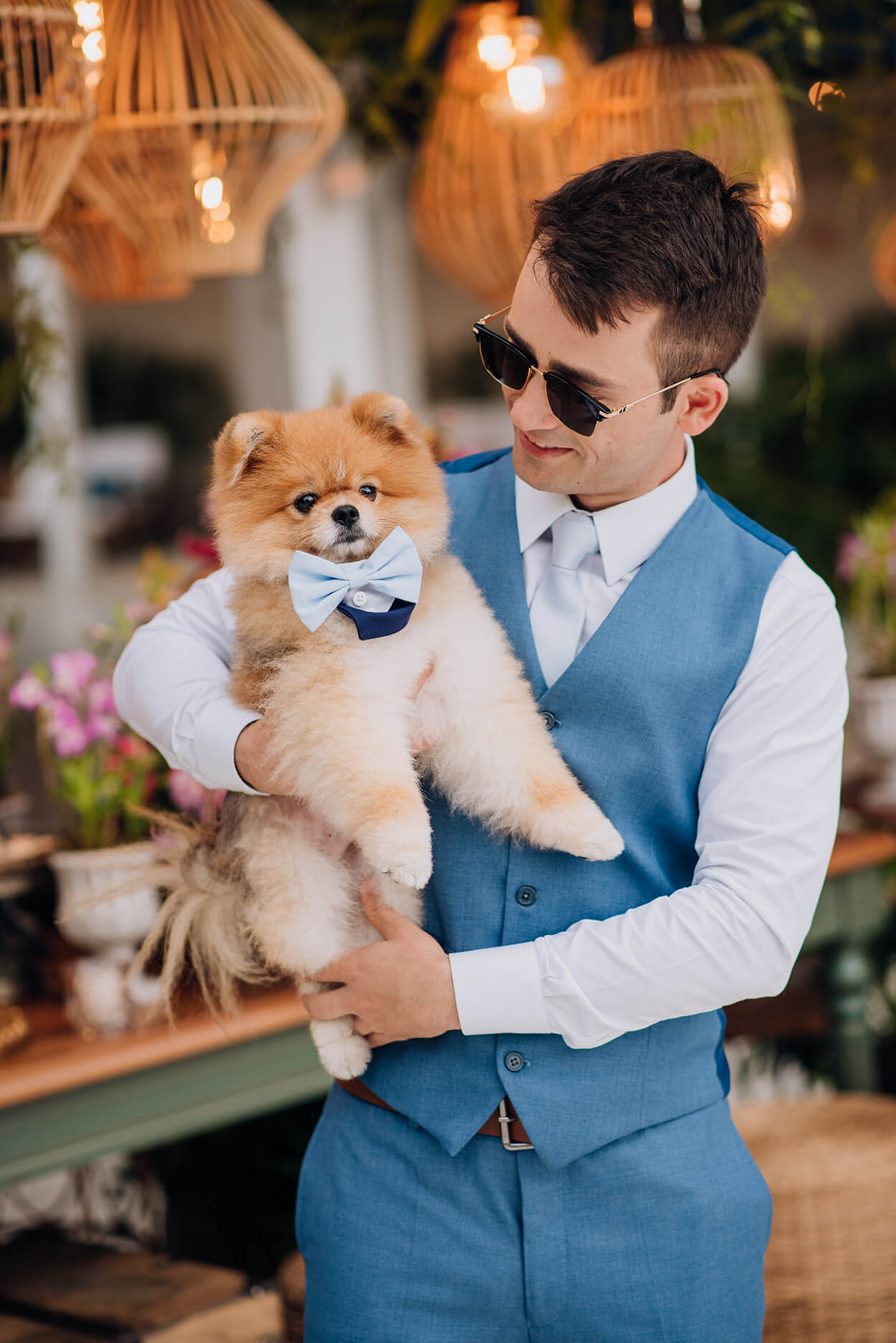 Noivo com camisa branca e colete azul segurando seu cachorro com gravata borboleta