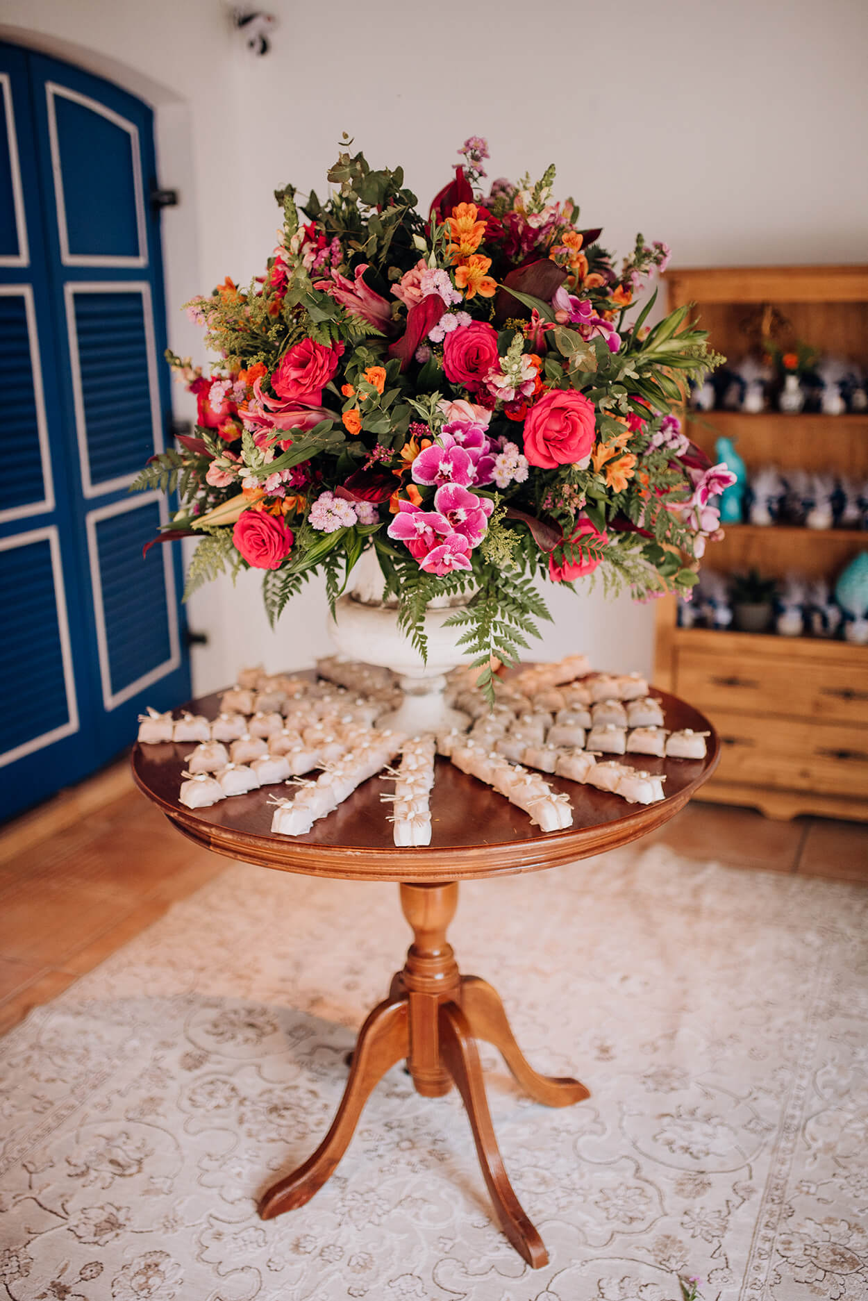 Mesa de madeira redonda com arranjo de flores vermelhas rosas e laranjas cercado por bem-casados