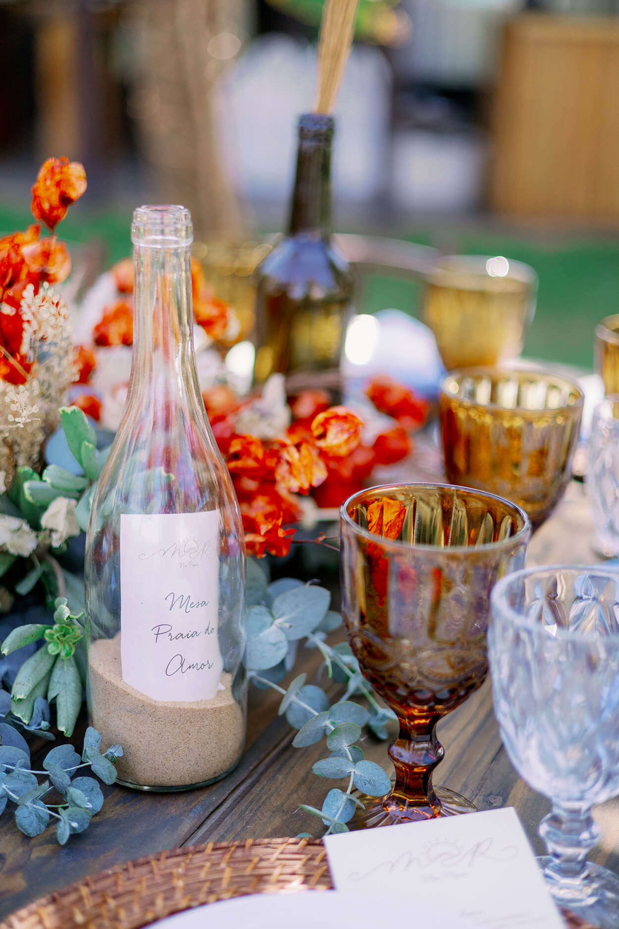 Centro de mesa com folhas de eucalipto e flores laranja e garrafa com areia dentro