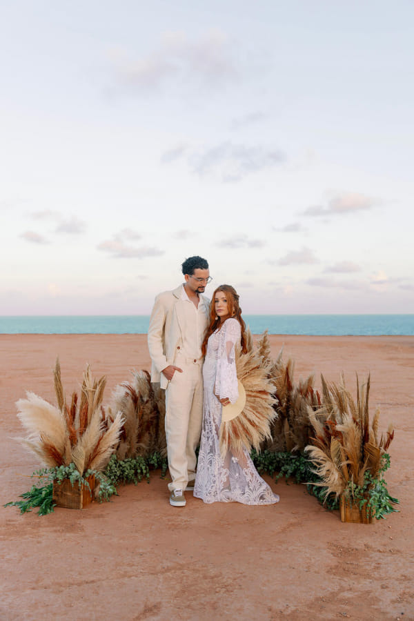 capa post: Noivo com terno de linho algodão cru de mãos dadas com a noiva com vestido longo segurando leque com capim dos pampas no altar com capim dos pampas