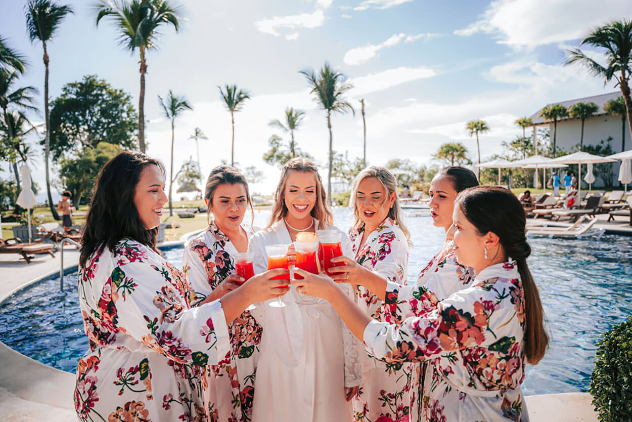 Noiva e madrinahs com robes floridos brindando drink na frente da piscina
