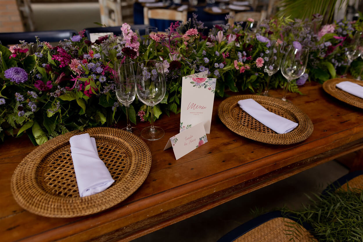 Mesa de madeira com sousplat rustico e flores rosas e lilas no centro da mesa