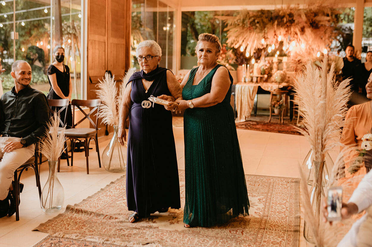 Duas senhoras com vestidos longos levando as alianças para os noivos