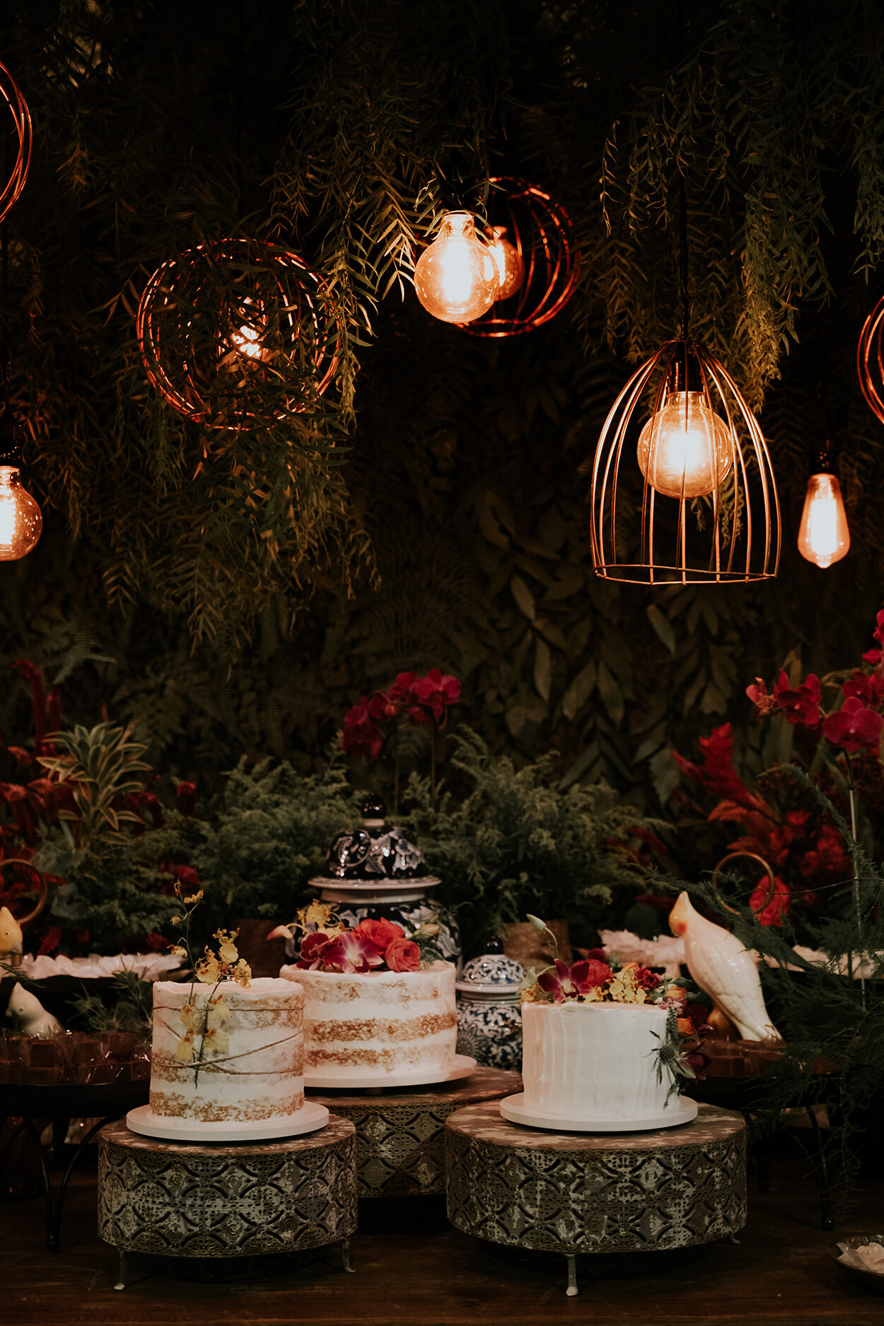 Mesa com flores vermelhas folhagens e trio de bolos de casamento brancos com topo com flores