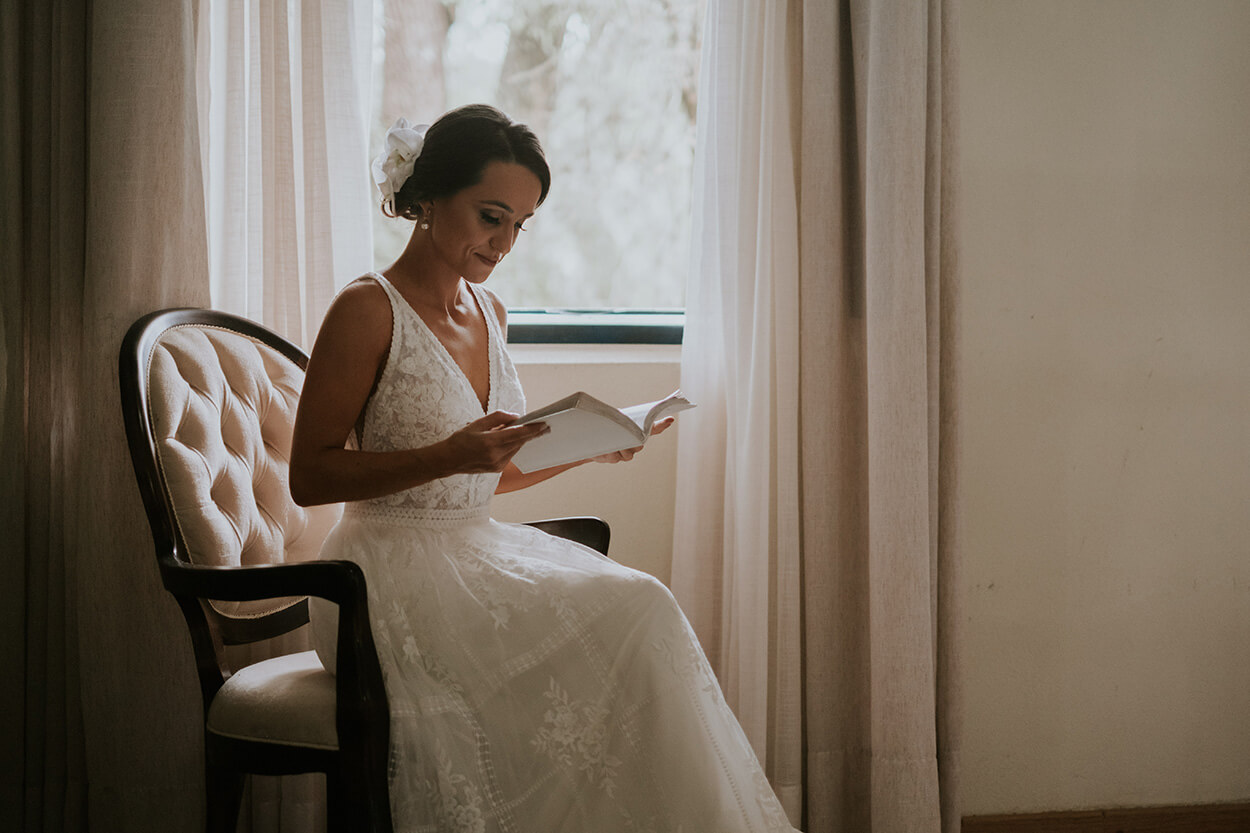 Mulher com vestido de noiva sentada na poltrona branca lendo caderno 