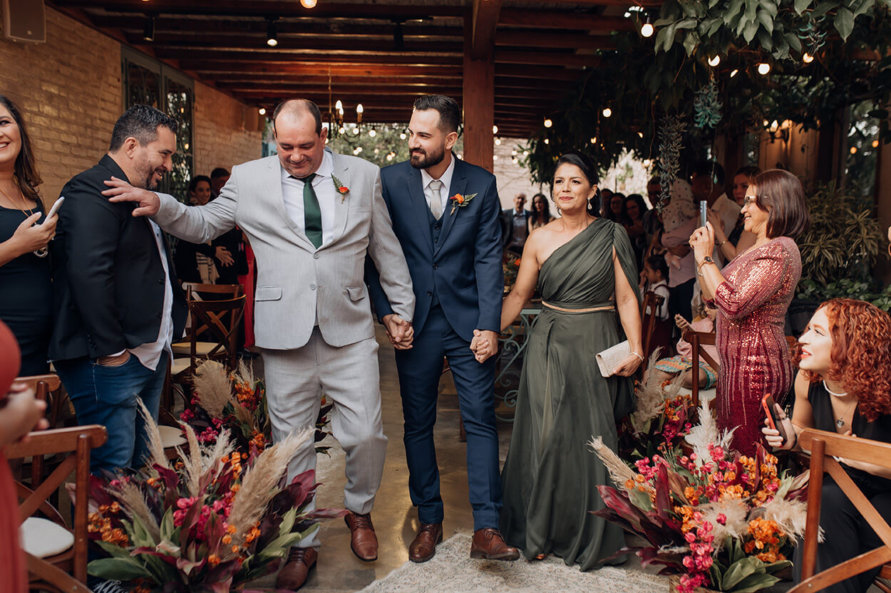 Entrada do noivo com terno azul ao lado do pai com terno cinza e gravata verde e ao lado da mãe com vetsido verde