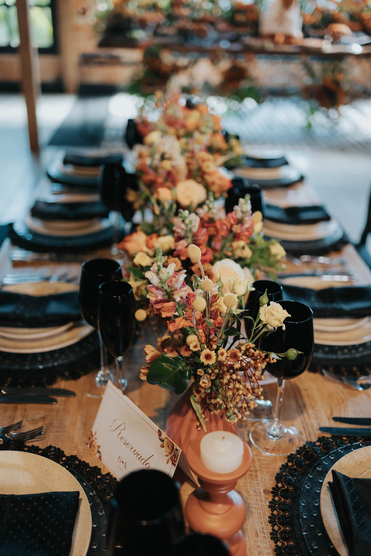 mesa posta com sousplat taça e guardanapo preto e flores amarelas e laranjas no centro