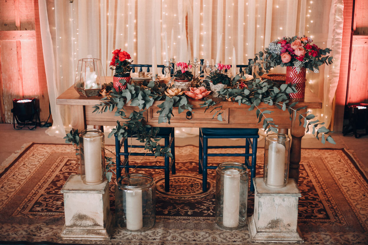 Mesa de madeira com flores vermelhas e laranjas e ramos de eucaliptos com cadeiras azuis e velas brancas dentro de vasos transparentes