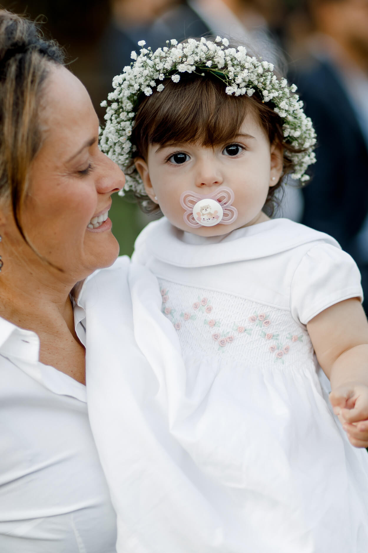 mulher carregando bebê daminha com coroa de flores brancas com chupeta