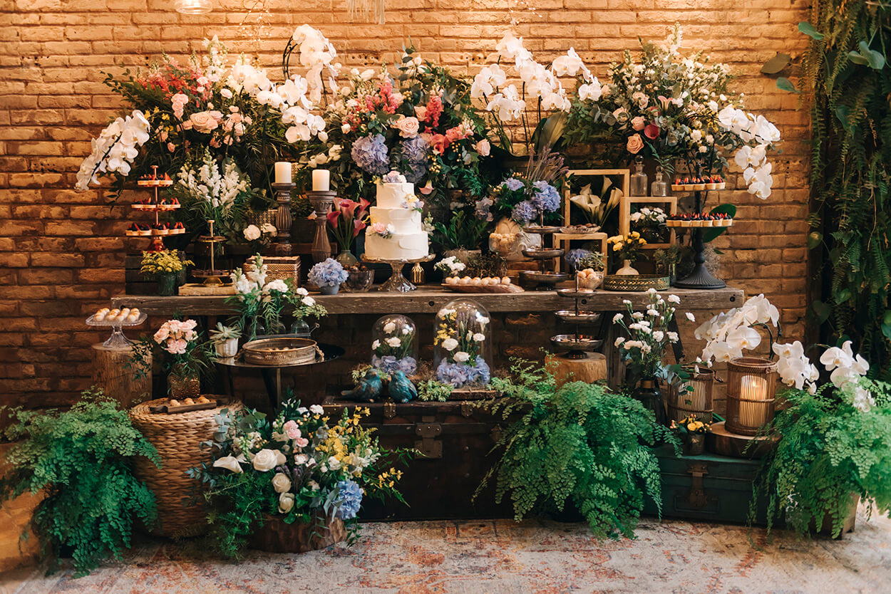 salão rústico com mesa do bolo com doces orquideas brancas hortensias azuis flores e folhagens