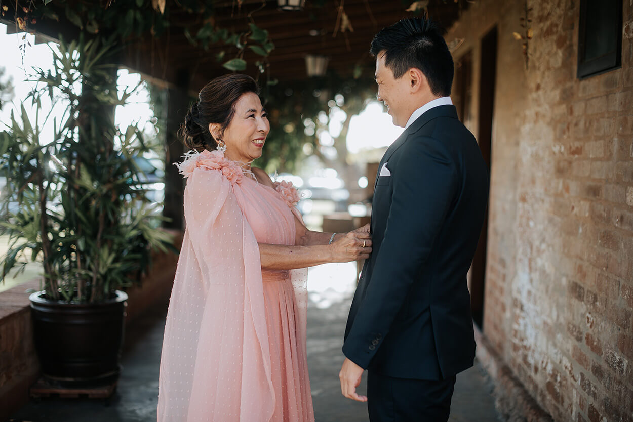 Mãe do noivo com vestido rosê com capa ajustando o terno do noivo