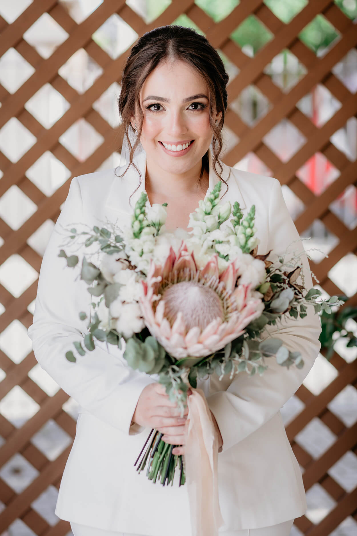 noiva com cabelo preso usando terno branco segurand buquê com flor protea