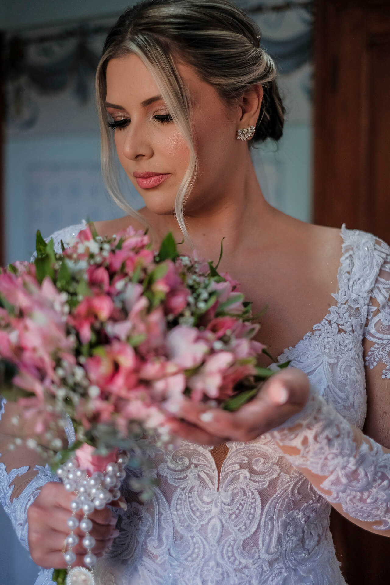noiva com cabelo preso olhando buquê com flores cor de rosa