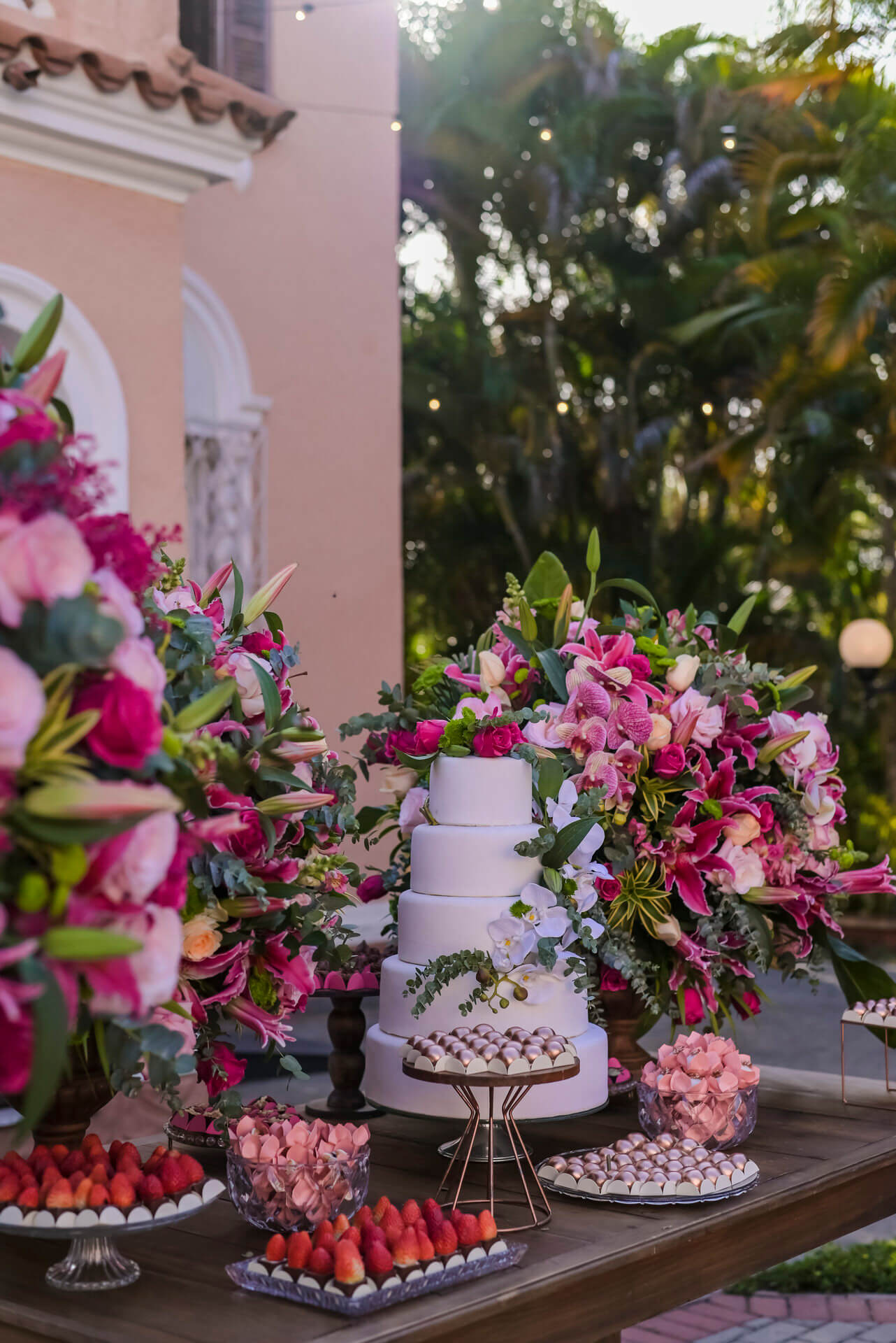 mesa de madeira com bolo de casamento com topo de flores e vasos com flores cor de rosa
