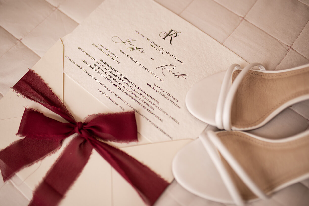Convite de casamento off white com laço marsala ao lado de sandalia branca