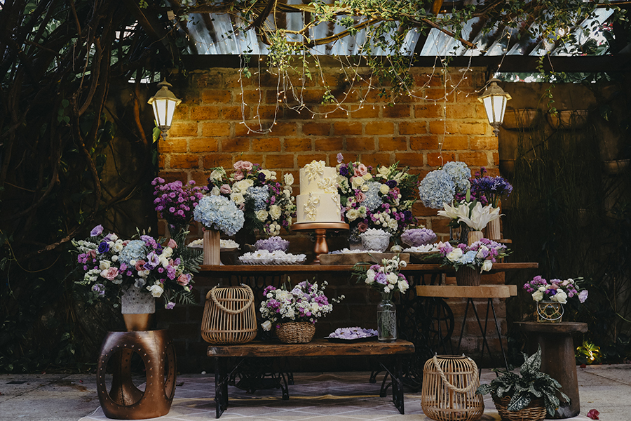 mesa com bolo de casamento branco e ao lado bandejas ocm doces finos e flores brancas azuis lavandas rosas 