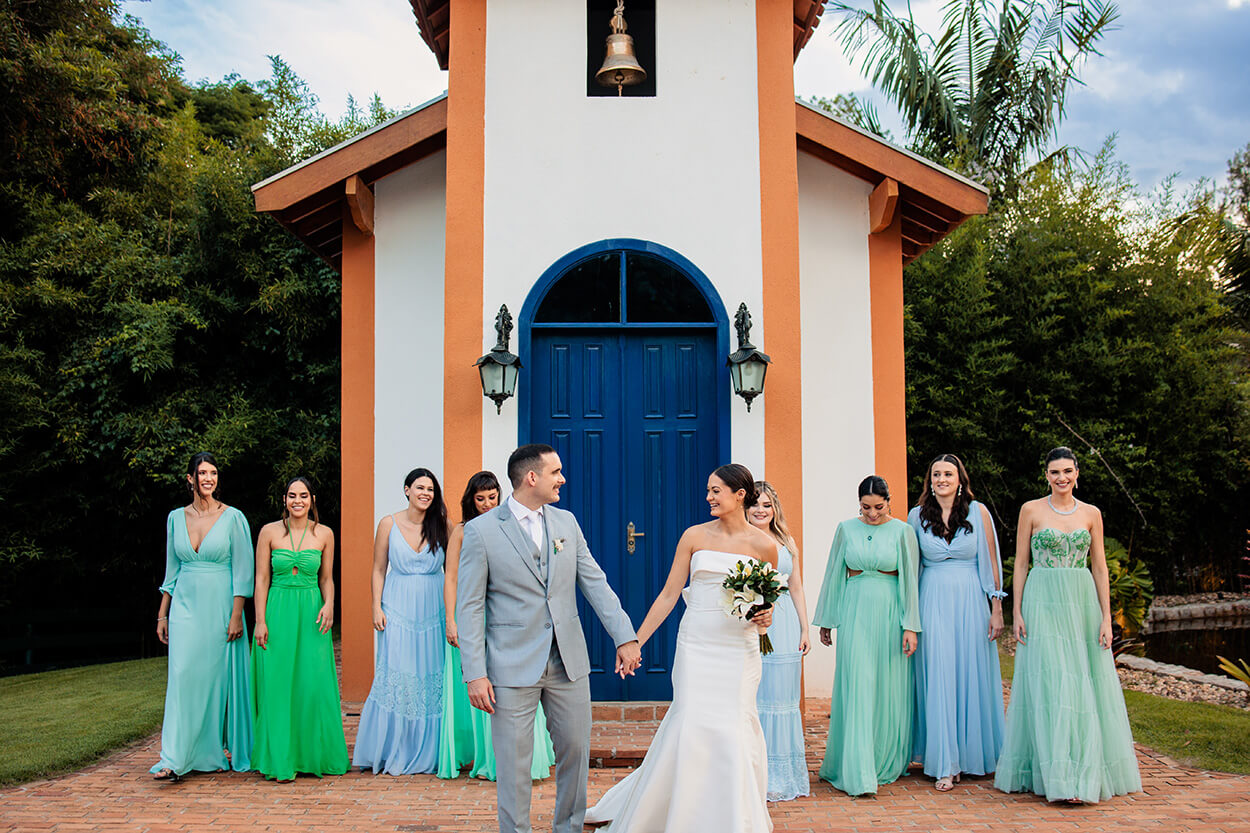 noivos de mãos dadas em frente a capela e madrinhas com vetsidos verdes claros e vestidos azul serenity