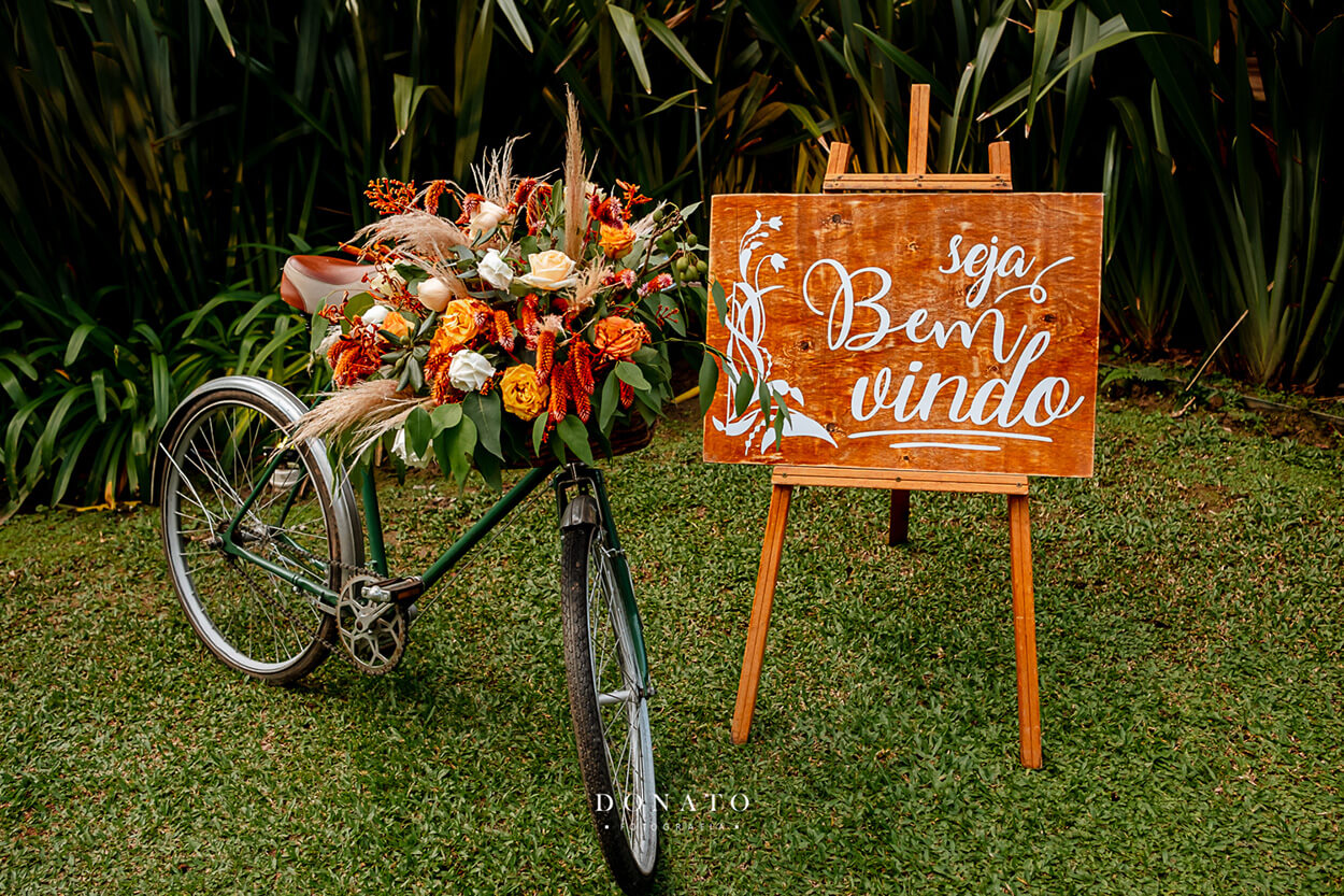 bicicleta verde com arranjo com flores brancas amarelas vermelhas e capim dos pampas ao lado de placa de madeira escrito bem vindos