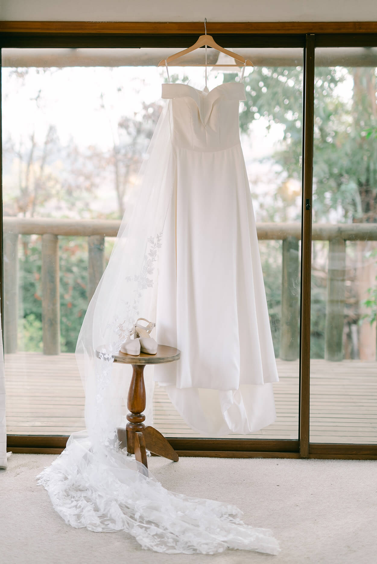 vestido de noiva pendurado no cabide ao lado do sapato branco da noiva em frente a janela