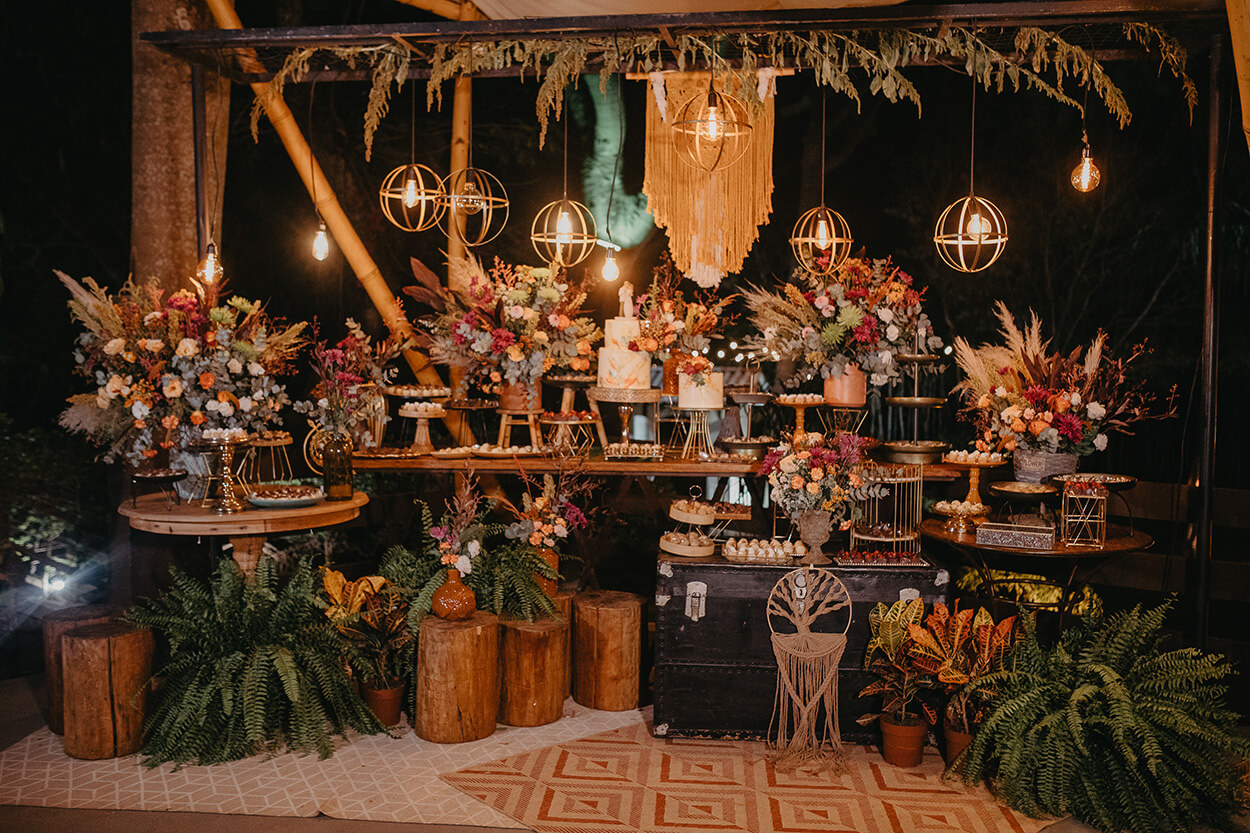 mesa de madeira com bolos de casamento com flores no topo ao lado de vasos de flores e bandejas com doces