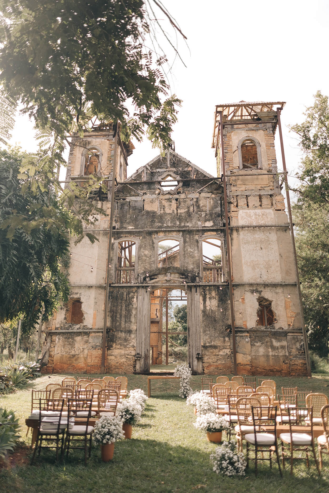Cadeiras de madeira e altar simples com flores brancas em jardim com ruínas de uma igreja
