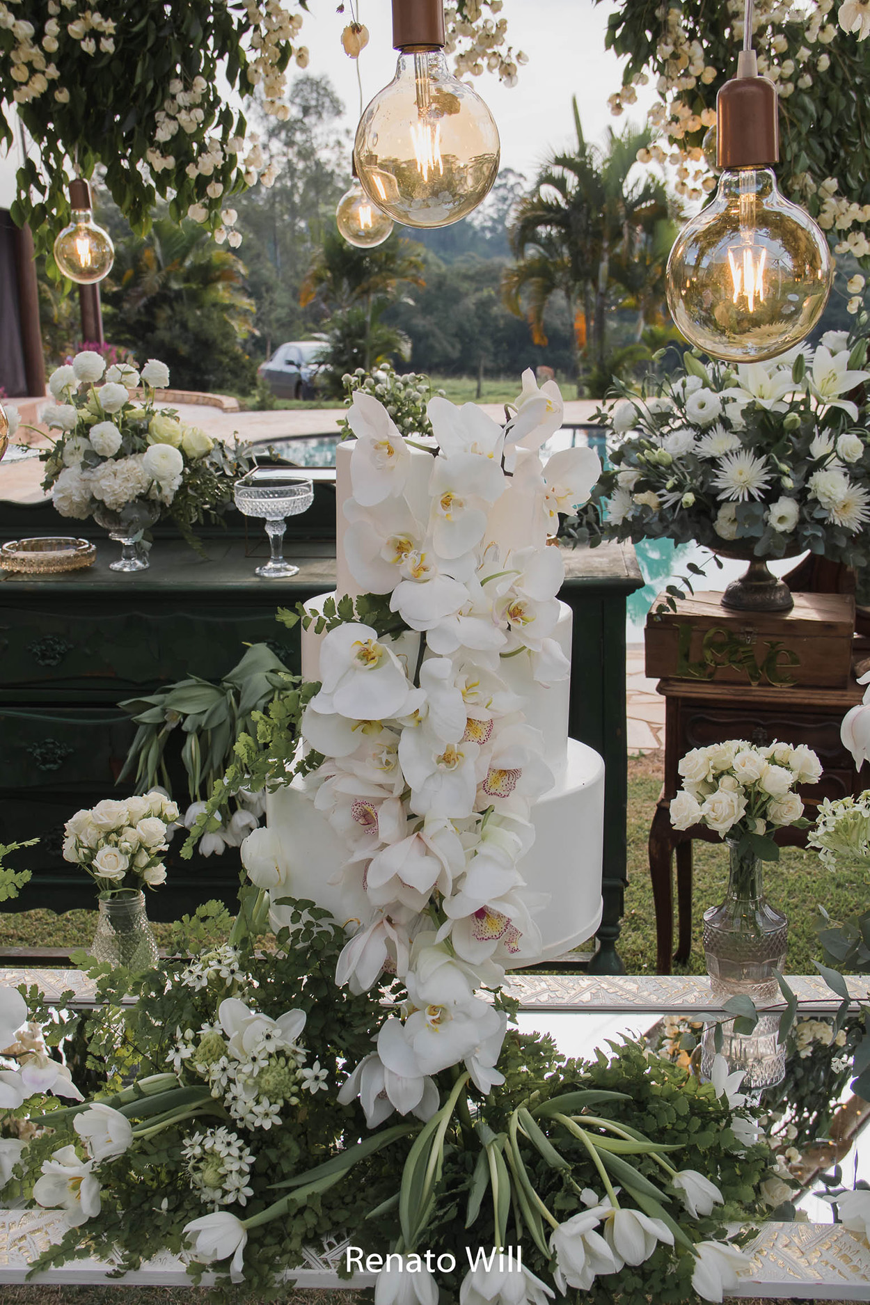 mesa com bolo de casamento branco com três andares com orquídeas brancas no topo