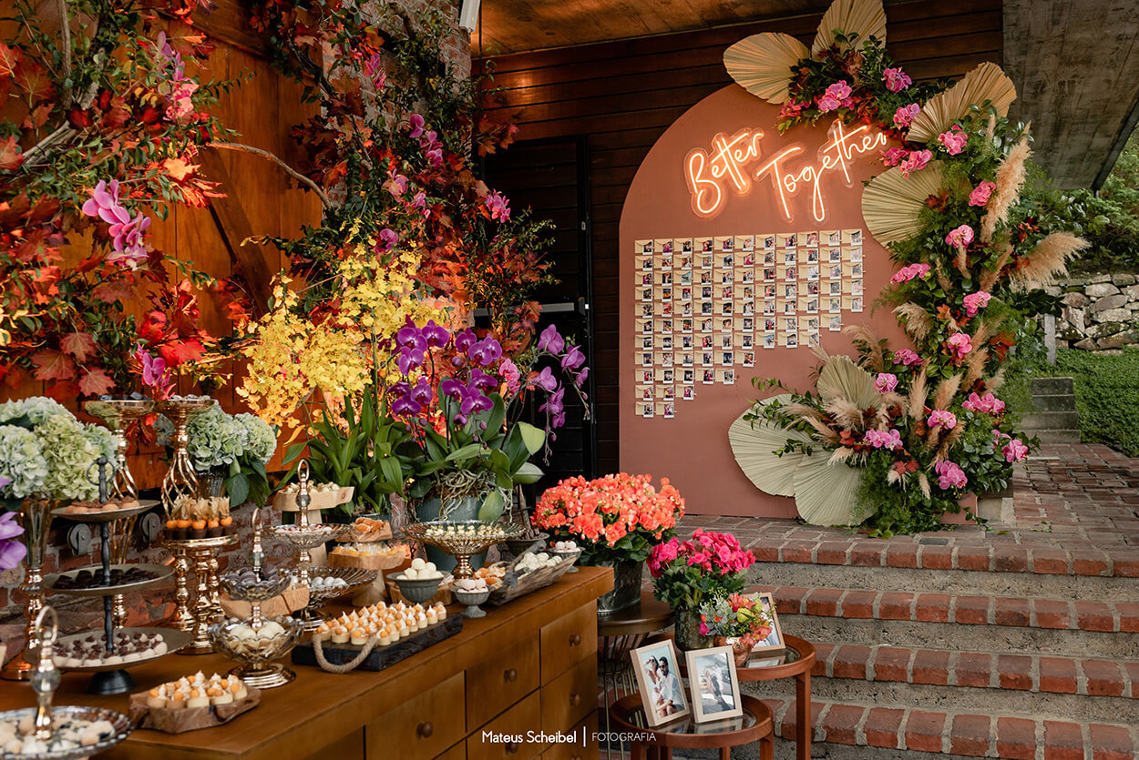 mesa com doces e vasos com flores ao lado de painel terracota com letreiro neon escrito better together e fotos