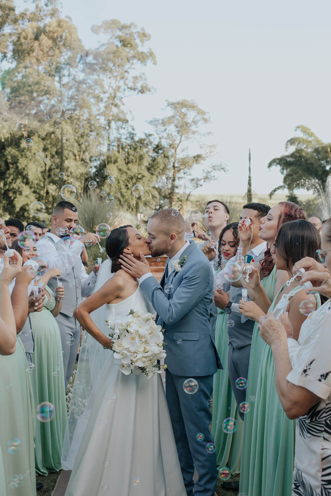 noivos se beijando durante a saída dos noivos ao ar livre e madrinhas e padrinhos soprando bolhas de sabão