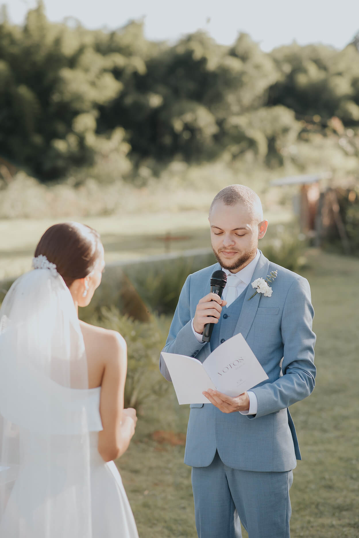 noivo com terno azul segurando o microfone e lendo os votos de casamento para a noiva ao ar livre
