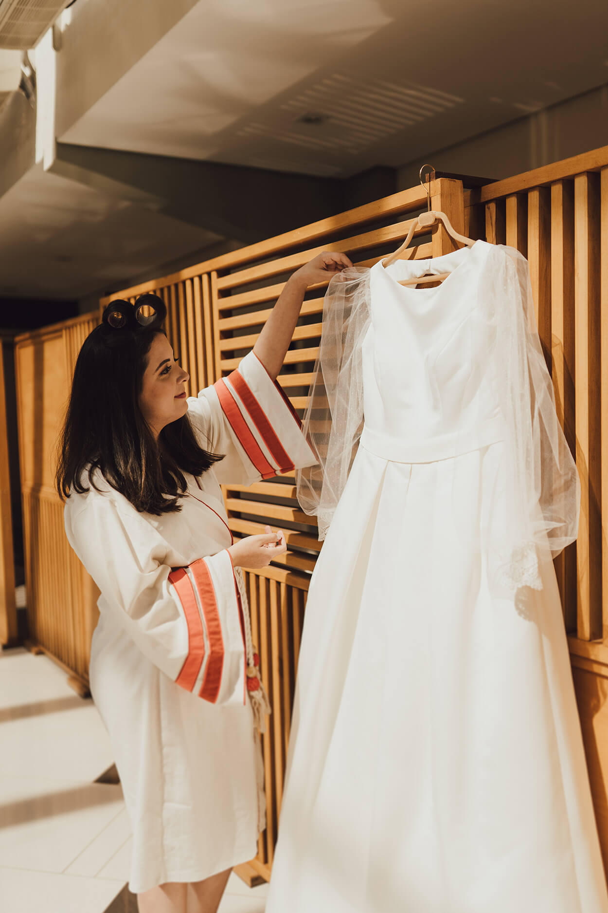 noiva com robe branco com detalhes de faixas laranjas na manga olhando o vestido de noiva com manga longa no cabide