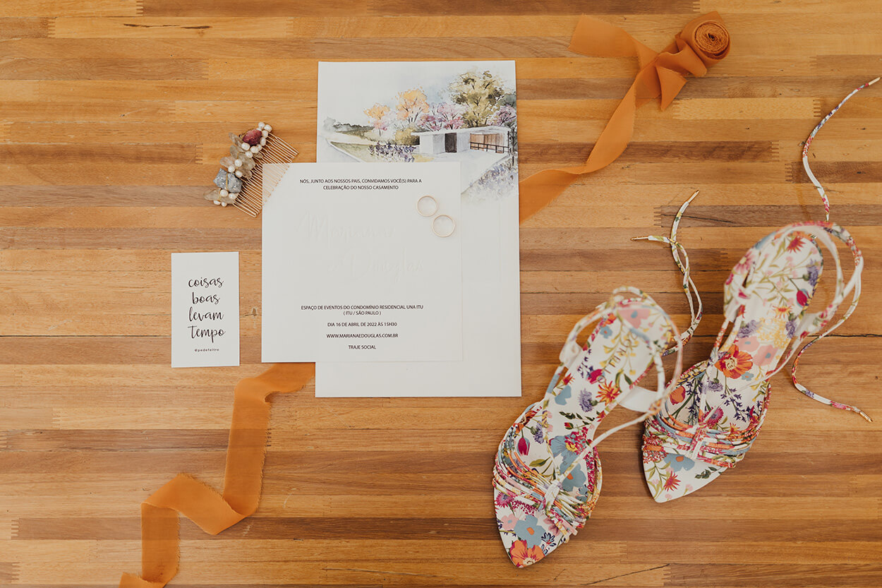 sandália floral estampada ao lado e convite de casamento com aquarela com ilustração de casa e fita terracota