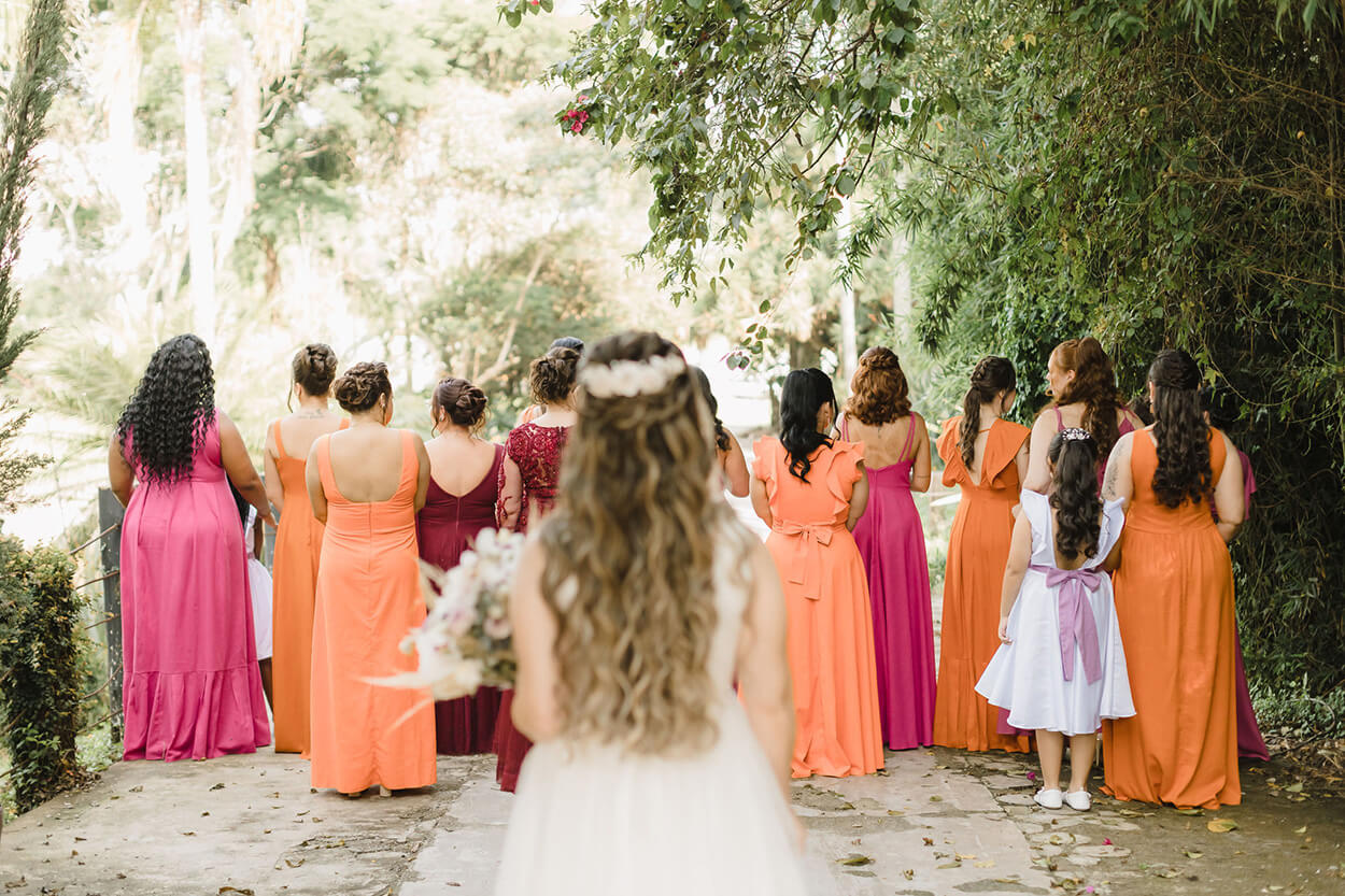 visão de noiva de costas com cabelo semi preso cacheado e mais a frente madrinhas com vestidos rosas e laranjas de costas