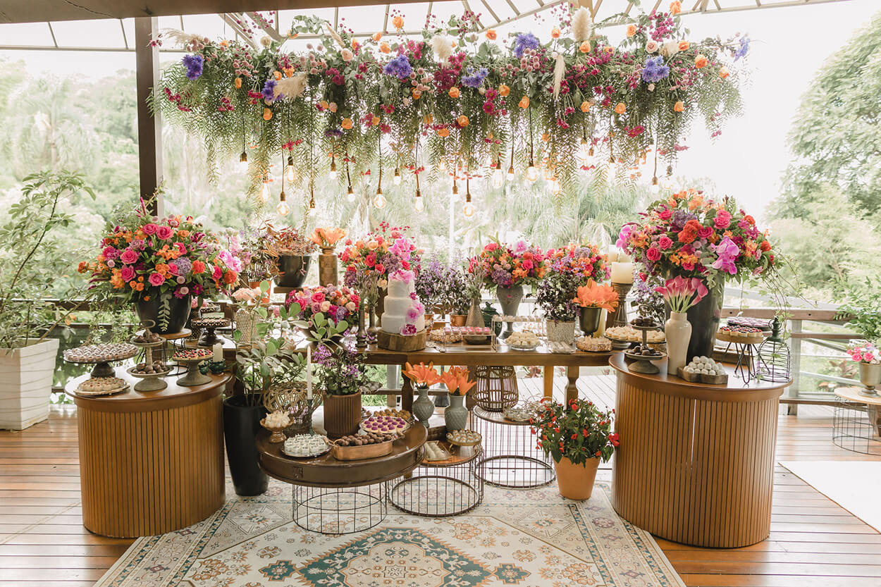 mesas de madeira com bolo de casamento branco com flores e vasos com flores rosas laranjas e lilas