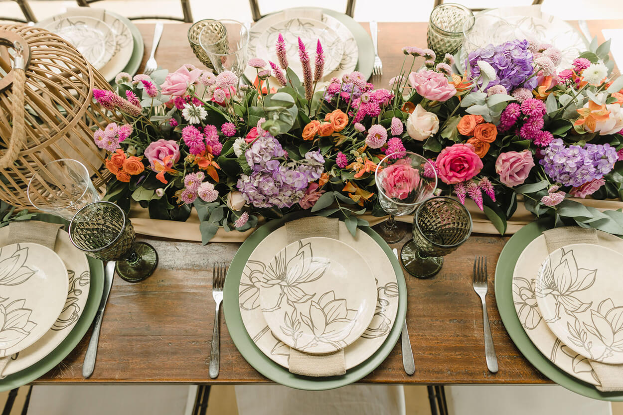 mesa de madeira com mesa posta com sousplat verde e flores rosas laranjas e lavanda no centro