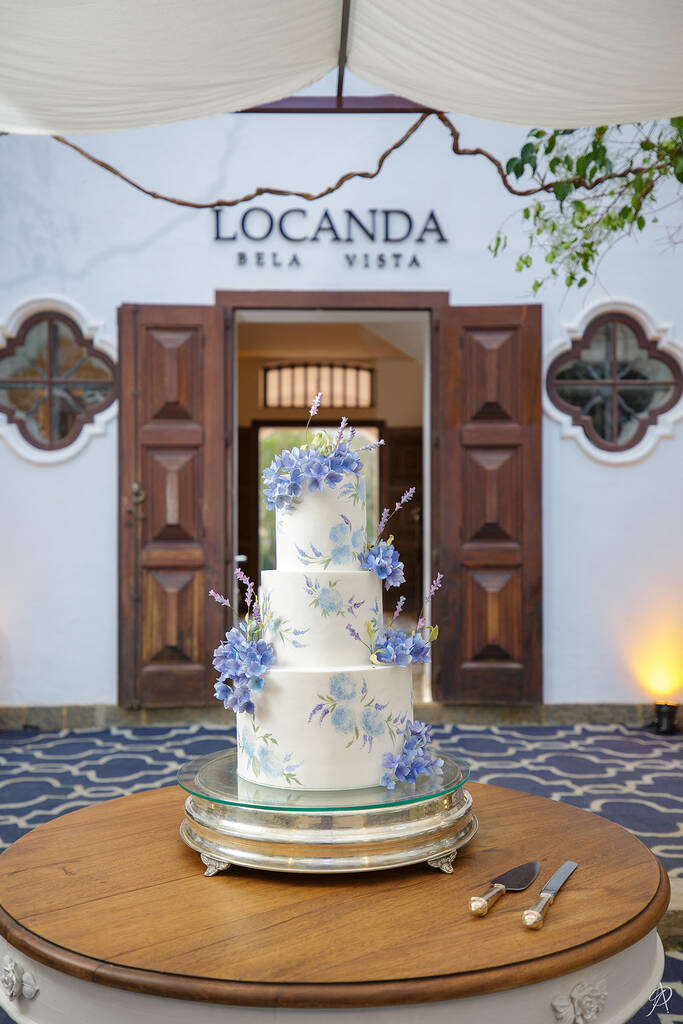 bolo de casamneto branco de três andares com flores azuis no topo e flores estampadas no bolo