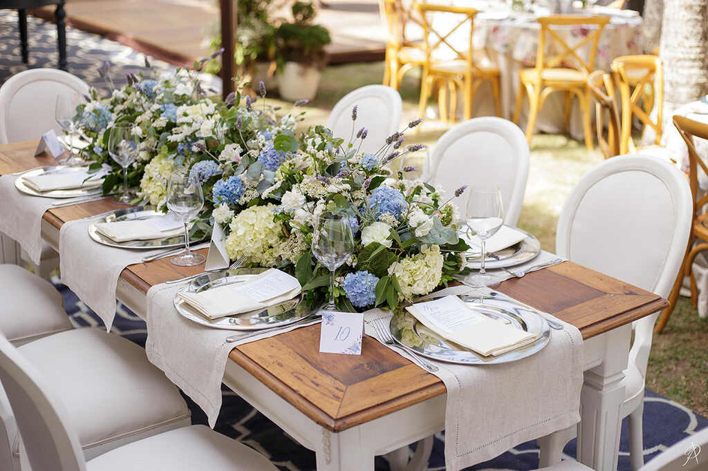 mesa de madeira posta com sousplat prateado com guardanapo branco e flores brancas e azuis no centro