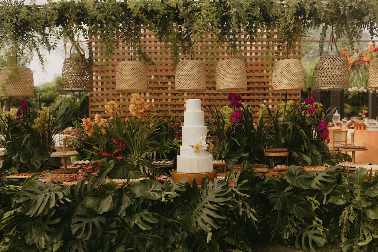 mesa com bolo de casamento branco moderno com cinco andares ao lado de vasos de flores com orquideas rosas e laranjas 