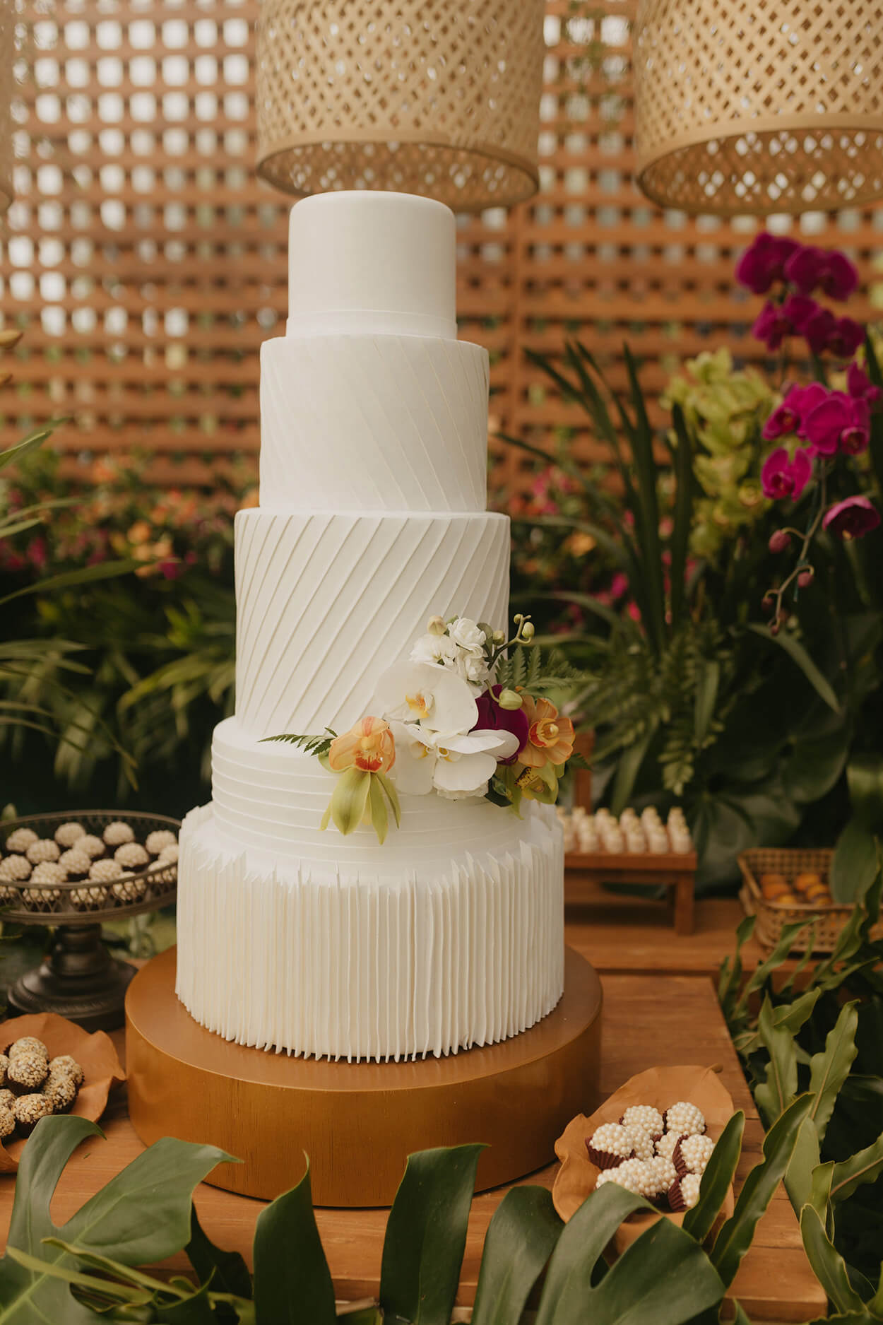 mesa com bolo de casamento branco moderno com cinco andares ao lado de vasos de flores com orquideas rosas e laranjas