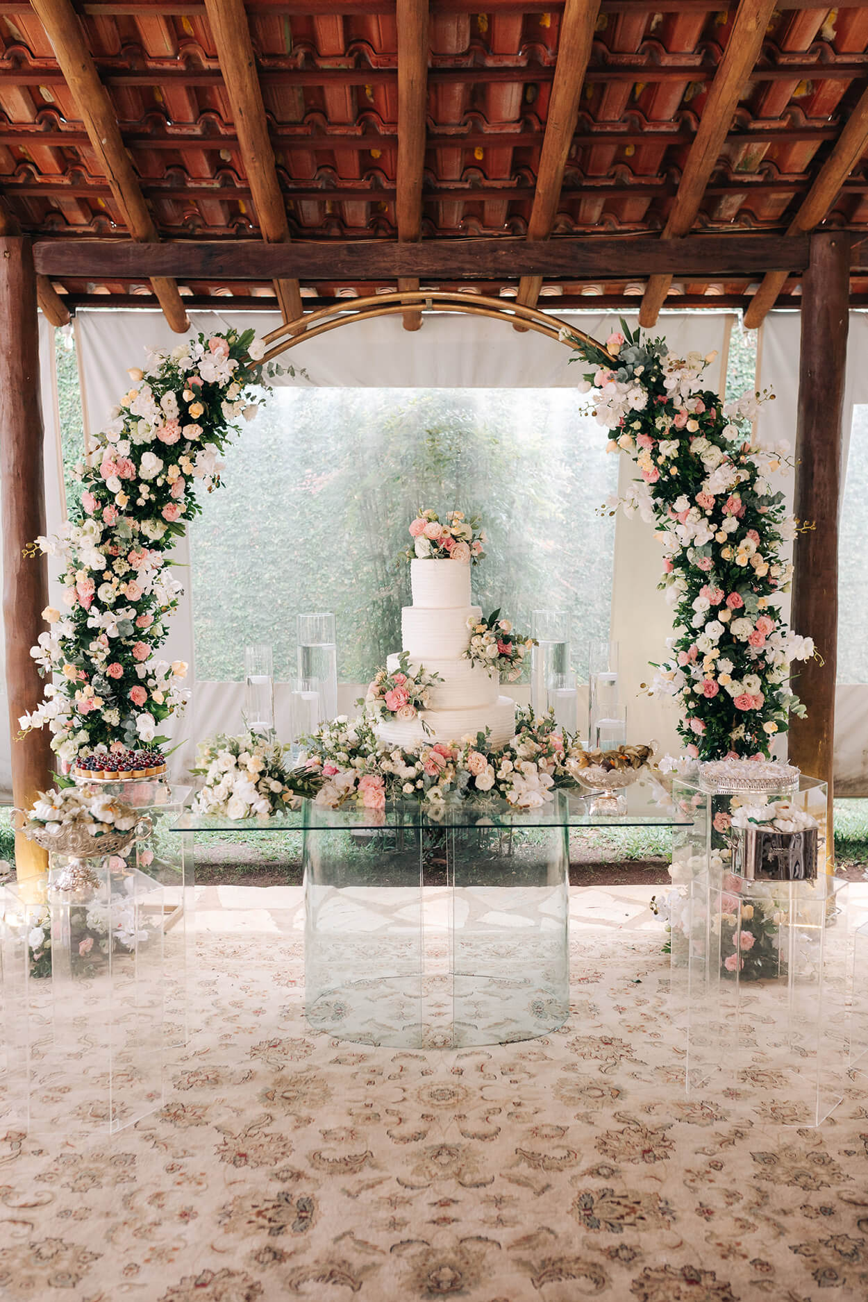 mesa de vidro com bolo de casamento branco com quatro andraes com flores amarelas brancas e rosas no fundo arco com flores brancas rosas e amarelas