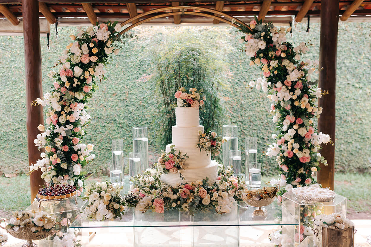 mesa de vidro com bolo de casamento branco com quatro andraes com flores amarelas brancas e rosas no fundo arco com flores brancas rosas e amarelas 